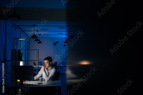 Bild auf Leinwand Remote view of serious young business man remote working overtime, learning online late at night in in dark room with neon light using desktop computer at workplace