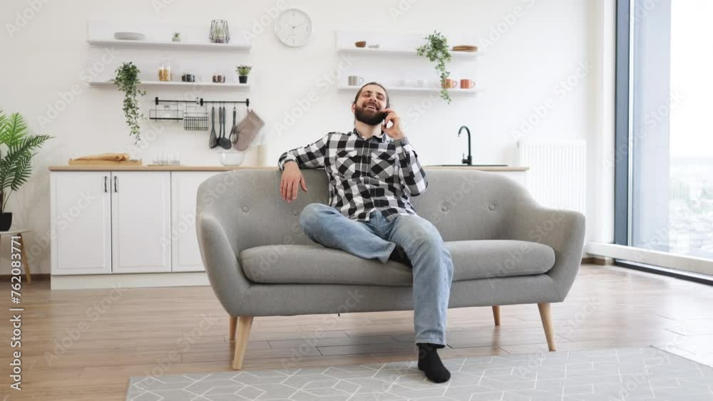 Efficient Caucasian freelancer having business conversation with client of remote company. Joyful young man in casual clothes talking on smartphone while sitting on sofa in spacious dining room. Efficient Caucasian freelancer having business conversation with client of remote company. Joyful young man in casual clothes talking on smartphone while sitting on sofa in spacious dining room.