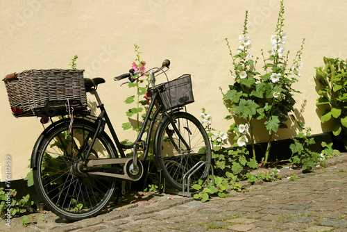 A city bicycle with two baskets, on the sidewalk, against a yellow wall.