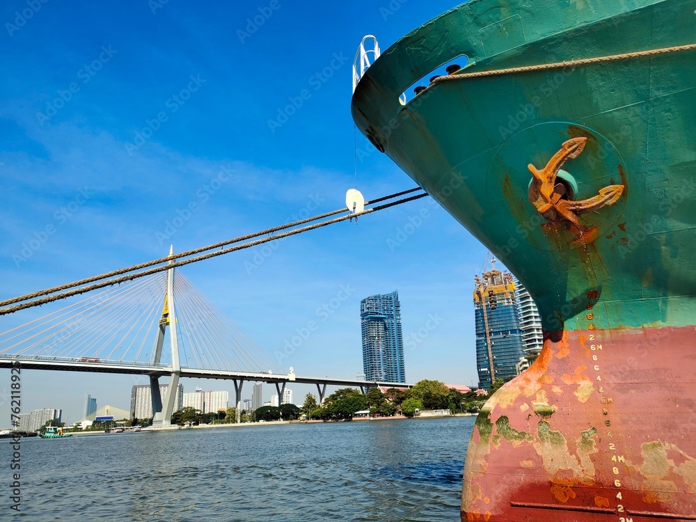 Fototapeta premium ship in the port, Anchor on large cargo ship's anchor being pulled. Green and red ship, While docked at the pier by large ropes on the river, the building background and transport concept