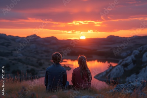 A serene image capturing a couple seated on grass overlooking a lake at sunset with the sky in vibrant hues of orange and pink © Jelena