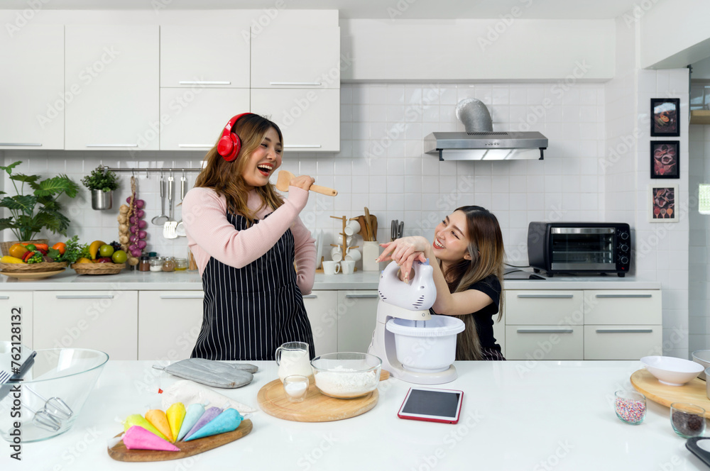Two women having a great time in a kitchen. Young asian woman with headphones and a sweater underneath an apron, sing while holding a wooden spatula. Her friend smile while listening to the music.