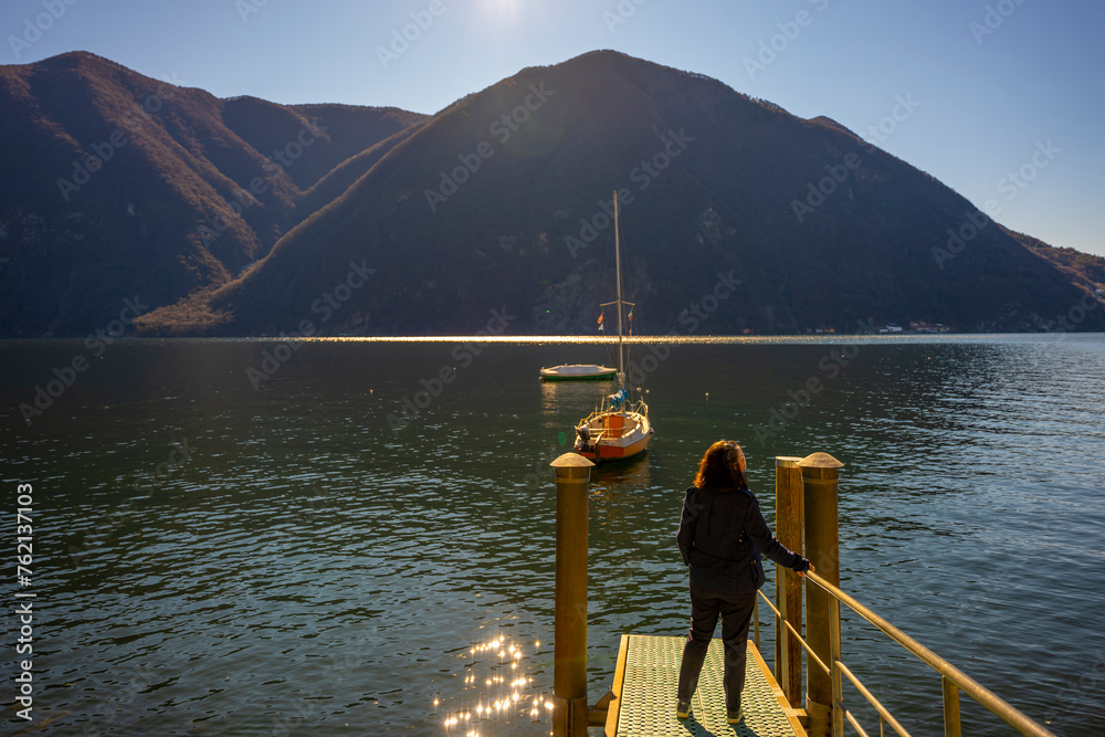Woman Standing on a Pier with Sailboat on Lake Lugano and Mountain Range in a Sunny Day with Clear Sky in Lugano, Ticino, Switzerland.