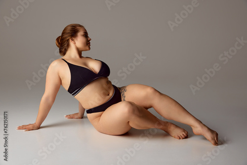 Portrait of young overweight woman posing in lingerie exuding elegance and self-contentment against grey studio background. Concept of beauty, femininity, body positivity, dieting, spa, wellness.