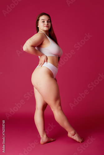 Young attractive, confident woman posing in while lingerie, bikini looking away against vibrant pink studio background. Concept of natural beauty, femininity, body positivity, dieting, fitness.