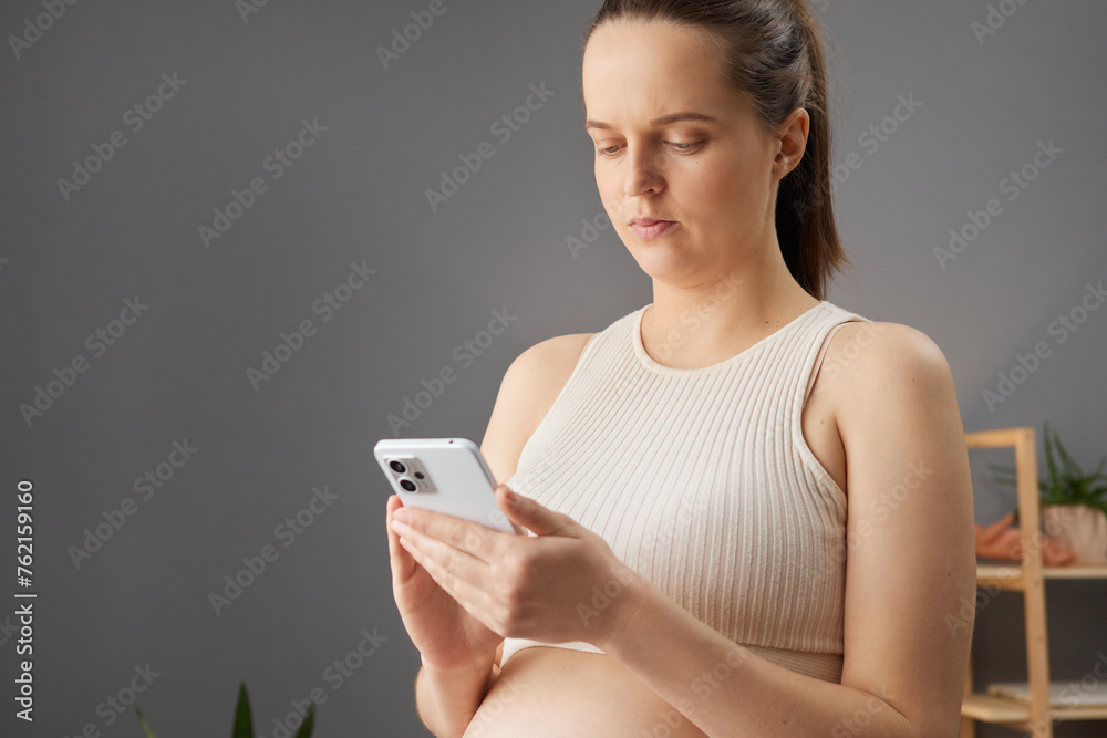 Serious brown haired pregnant woman in beige top using mobile phone browsing web pages scrolling online looking at device screen chatting with doctor gynecologist Serious brown haired pregnant woman in beige top using mobile phone browsing web pages scrolling online looking at device screen chatting with doctor gynecologist