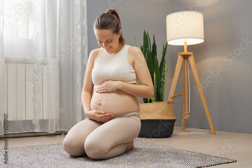 Adorable pleased pregnant woman wearing beige clothing sitting on floor in living room touching and looking at her tummy restring at home enjoying pregnancy time photo
