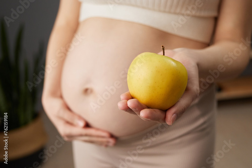 Unknown beautiful pregnant woman with bare belly standing in home interior holding fresh organic yellow apple prefers healthy eating while expecting baby photo