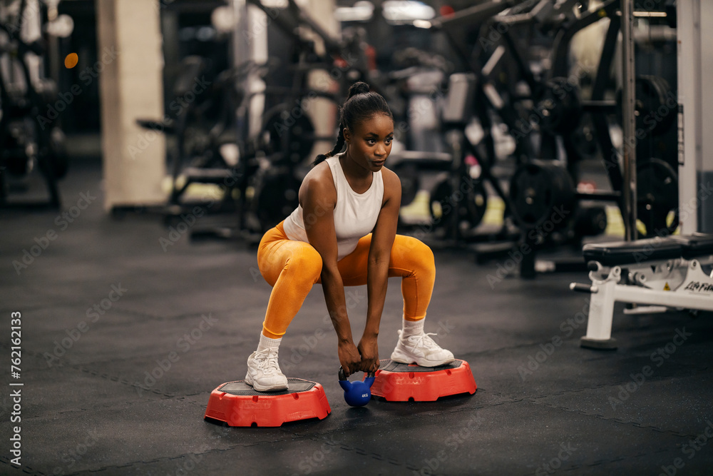 A strong black sportswoman doing deep squats with kettlebell at gym. - obrazy, fototapety, plakaty 