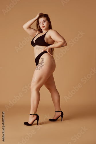 Full length portrait of young chubby woman posing in lingerie standing on heels against sandy color studio background. Self-expression. Concept of natural beauty, femininity, body positivity, spa.