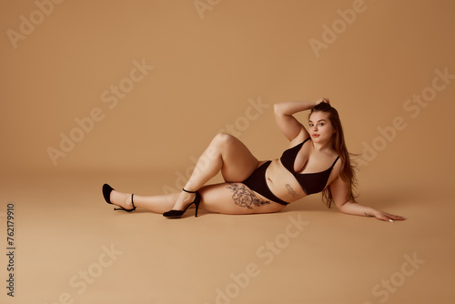 Portrait of young overweight woman posing in lingerie and heels shows elegance and self-contentment against beige studio background. Concept of natural beauty, femininity, body positivity, diet, spa.