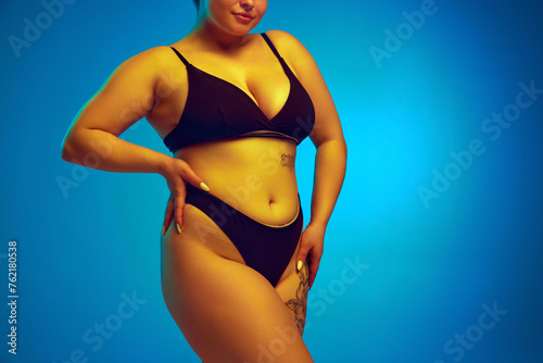 Woman, model plus size in dark lingerie, showcasing natural body curves in yellow neon light against blue studio background. Concept of natural beauty, femininity, body positivity, dieting, fitness. © Lustre