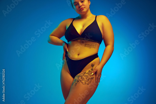Cropped portrait of female plus-size model posing in dark bikini, underwear in yellow neon light against blue studio background. Concept of natural beauty, femininity, body positivity, diet, fitness. © Lustre