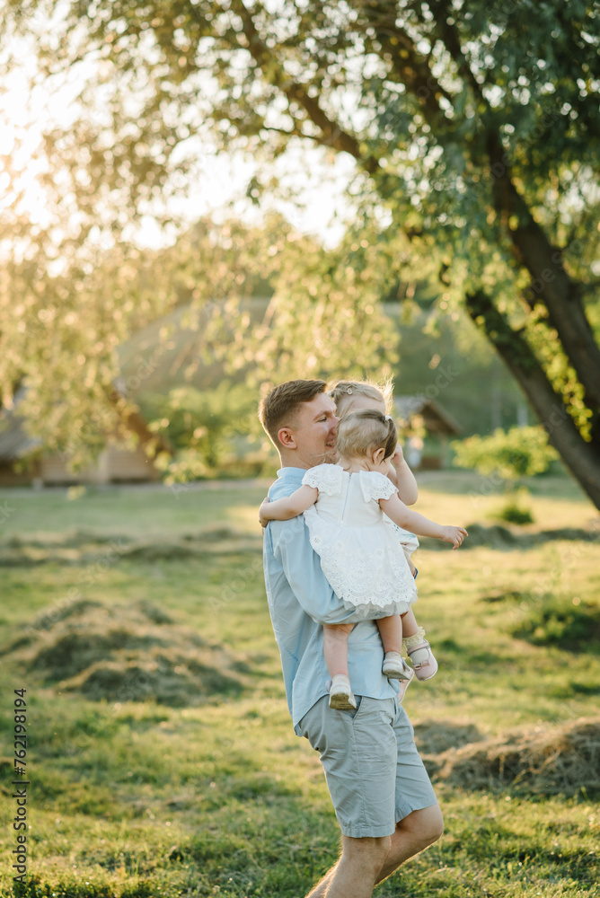 Happy dad embraces child's girls in park at sunset. Daddy hugs kids and spends time together. Father with two daughters spring day. Portrait of a daughter hugging father in a field in summer. Closeup Happy dad embraces child's girls in park at sunset. Daddy hugs kids and spends time together. Father with two daughters spring day. Portrait of a daughter hugging father in a field in summer. Closeup