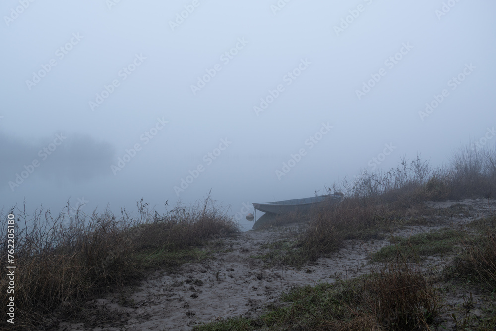 Muddy path leads to moored boat in fog, calm foggy evening near river, nautical vessel