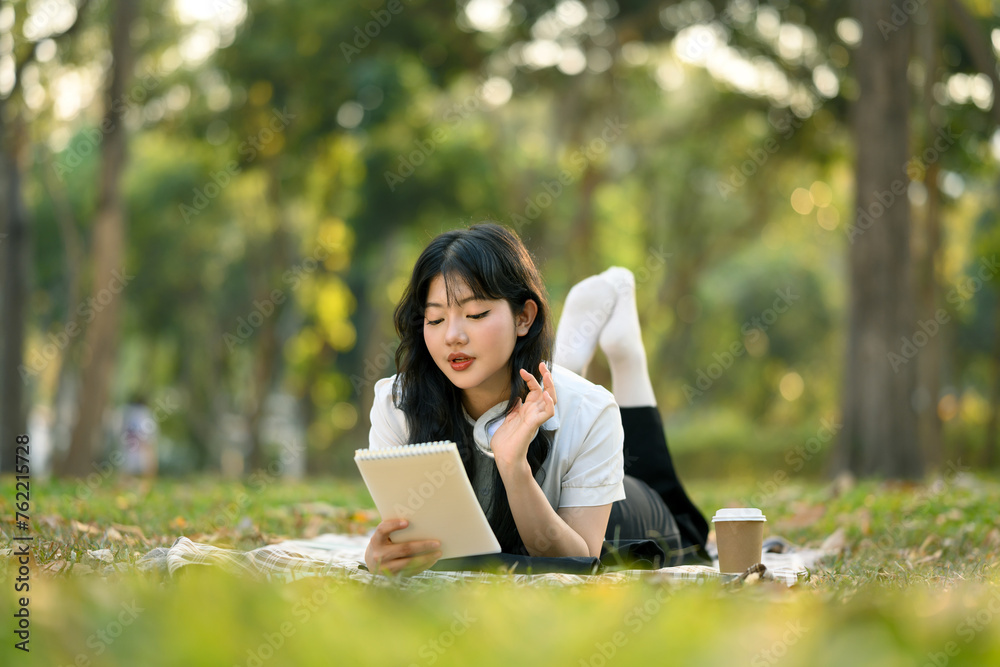 Calm Asian girl in casual clothes lying on picnic blanket and reading notes in notebook Calm Asian girl in casual clothes lying on picnic blanket and reading notes in notebook