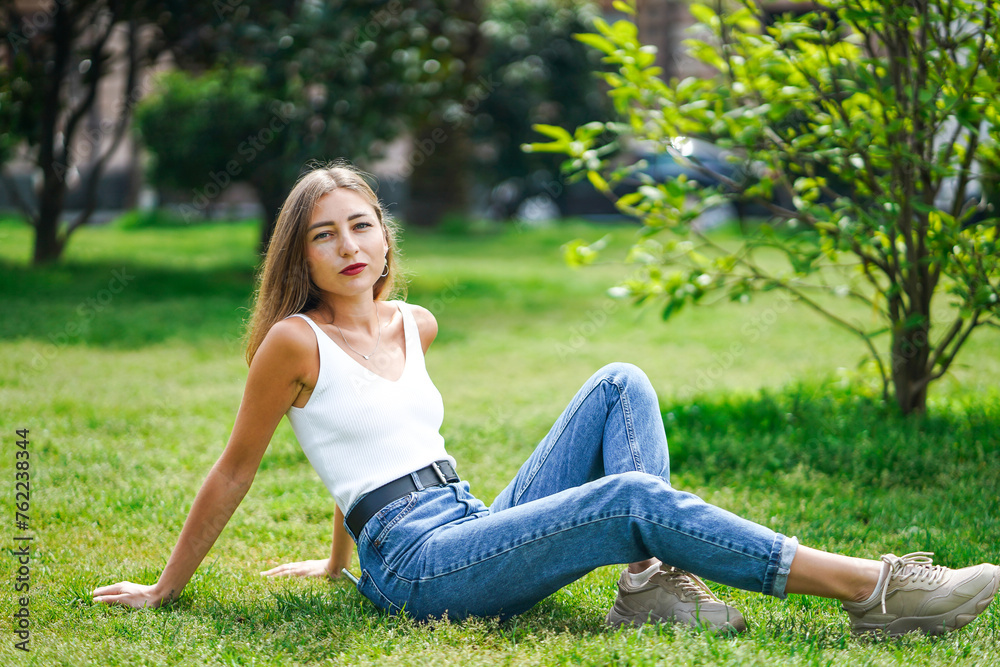 Beautiful young woman wearing tank top and jeans sitting on grass lawn Beautiful young woman wearing tank top and jeans sitting on grass lawn