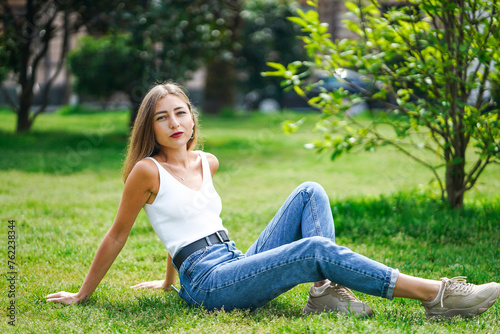 Beautiful young woman wearing tank top and jeans sitting on grass lawn