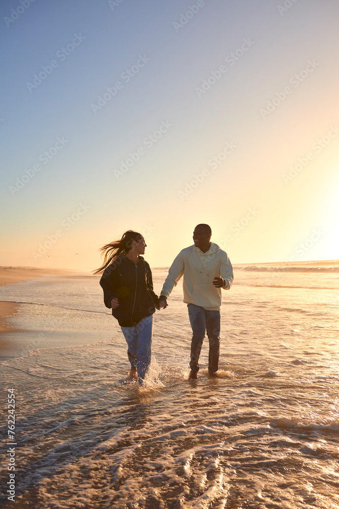 Casually Dressed Loving Young Couple Running Along Beach Shoreline Through Waves At Sunrise