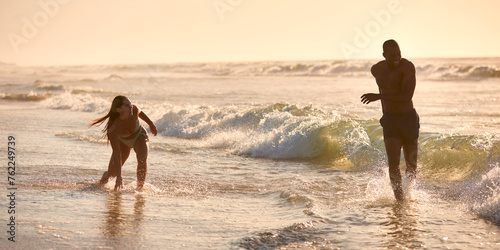Young Couple Wearing Swimwear On Vacation Running Along Beach Shoreline Through Waves