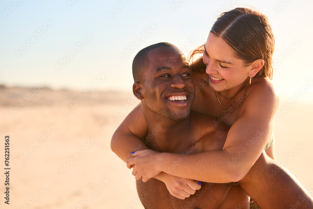 Loving Couple Wearing Swimwear With Man Giving Woman Piggyback Ride On Beach Shoreline Loving Couple Wearing Swimwear With Man Giving Woman Piggyback Ride On Beach Shoreline