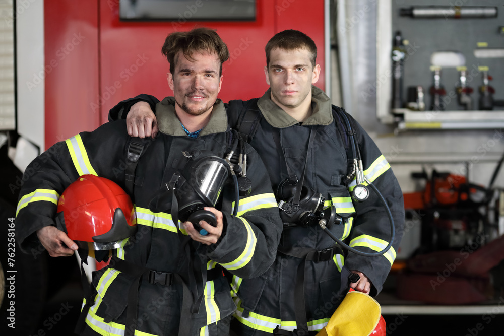 Two firefighters in protective suits standing against fire engine, one hugs other by shoulder Two firefighters in protective suits standing against fire engine, one hugs other by shoulder