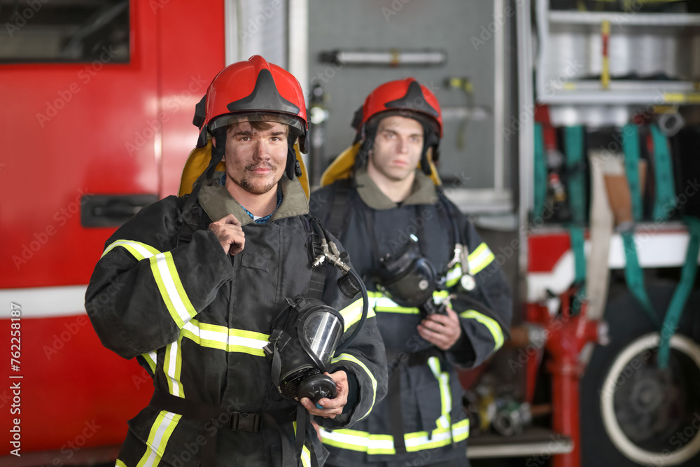 Two firefighters in protective suits and red fire helmet to protect head standing against fire engine Two firefighters in protective suits and red fire helmet to protect head standing against fire engine