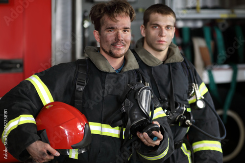 Two firefighters in protective suits standing against fire engine