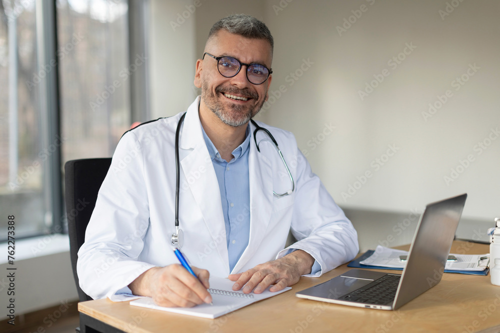 Happy middle aged man doctor in white medical coat, working at his workplace at modern clinic, writing prescription and using laptop computer, smiling at camera