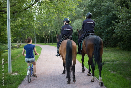 Back of two horse policemen and woman (model with release) rides bike in park at Sokolniki park.