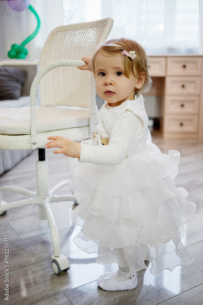 Little girl in white holiday dress stands holding hand on armrest of armchair. Little girl in white holiday dress stands holding hand on armrest of armchair.