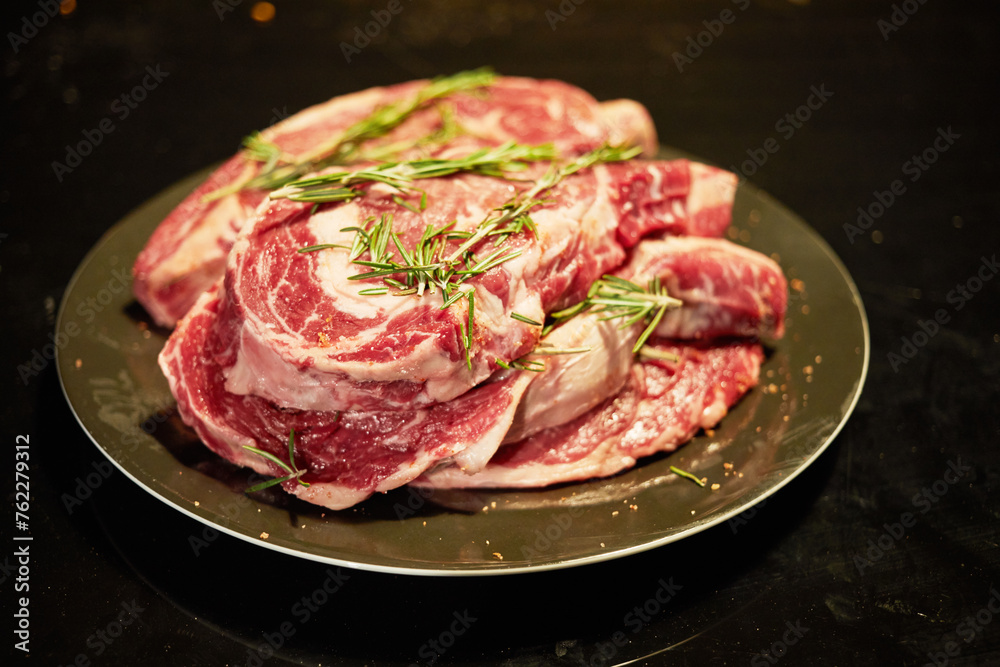 Pile of raw rib-eyes with rosemary and pepper on plate.