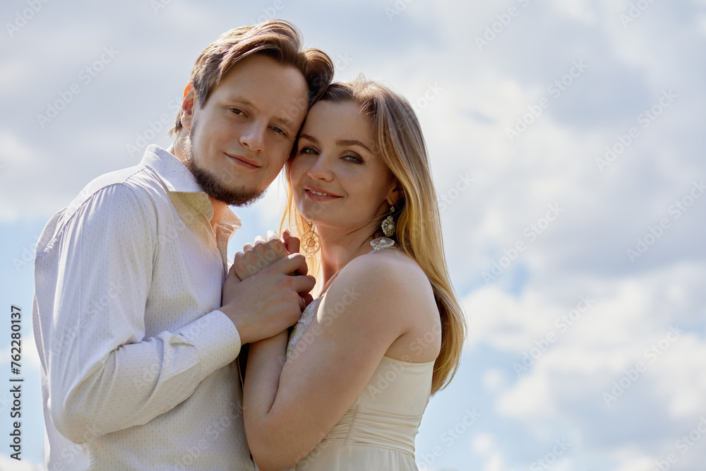 Young smiling bearded man and woman stand with their heads pressed together against cloudy sky.