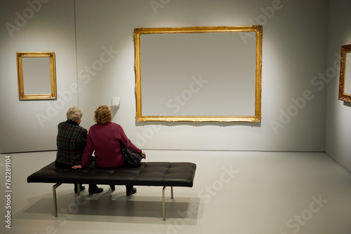 Two women sit on couch in room with frames on walls.