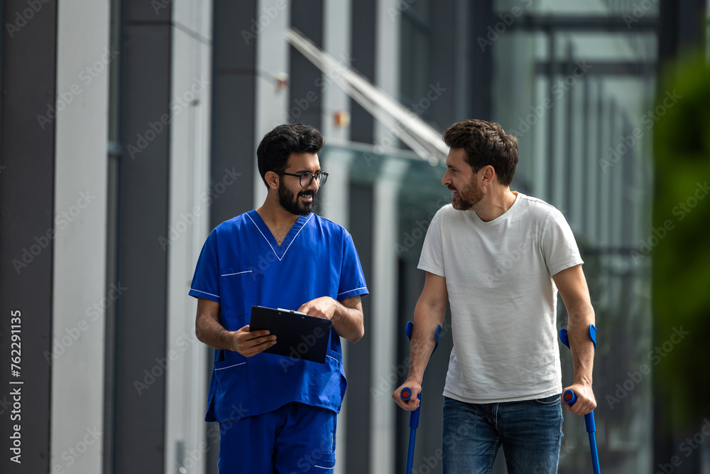 Young bearded man with crutches walking and talking to a male nurse Young bearded man with crutches walking and talking to a male nurse