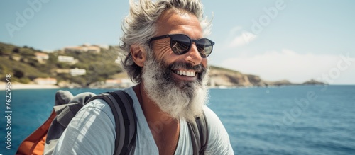 A smiling man with a beard and sunglasses is enjoying the sea breeze on a boat, with the blue sky above. His eyewear reflects a happy travel adventure