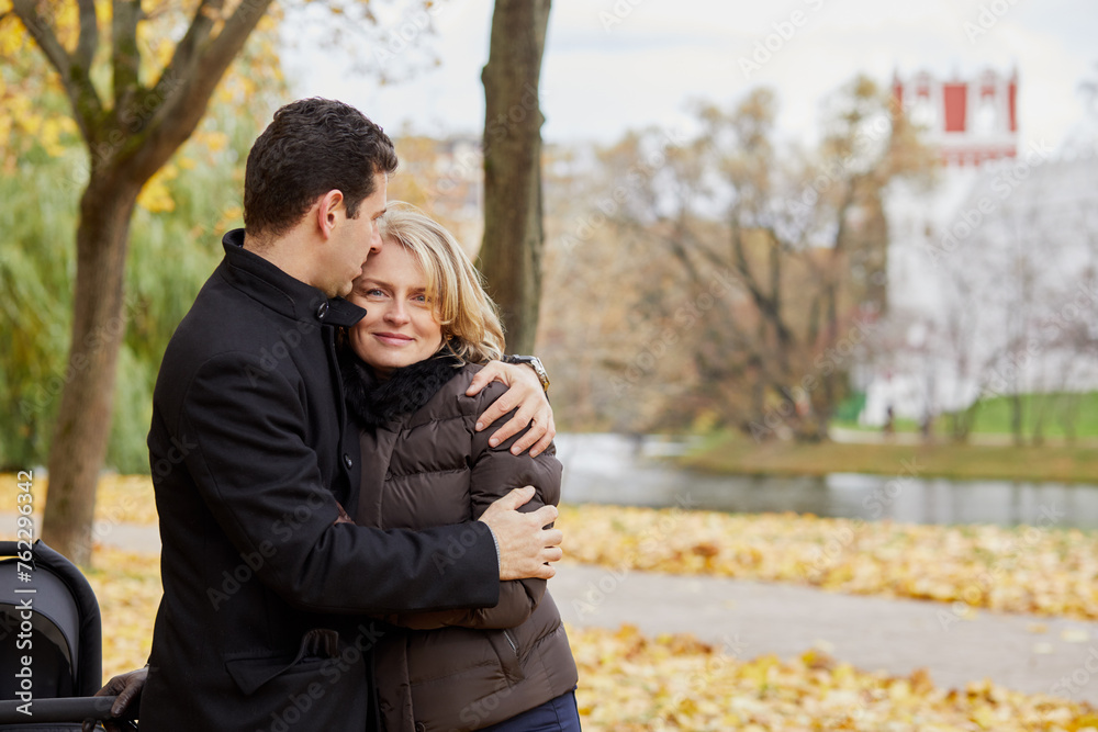 Man and woman with perambulator stand embracing in autumn park on river bank. Man and woman with perambulator stand embracing in autumn park on river bank.