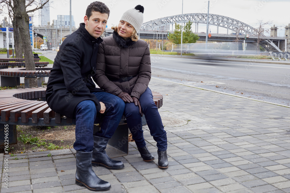 Man and woman sit on bench near city road on autumn day Man and woman sit on bench near city road on autumn day