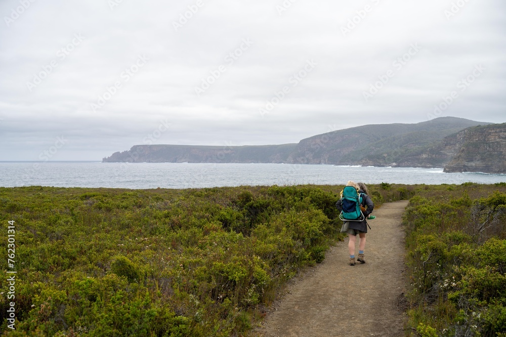 Woman and toddler hiking in the Tasmanian wilderness in the bush under the mountain and above the water camping in the wild with a backpack in Australia