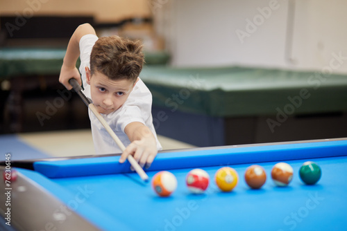 Boy in white polo-neck shirt aims ball with cue stick in billiard club.