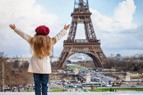 A happy tourist girl in a trench coat and with a red beret hat looks at the famous Eiffel Tower in Paris, France © moofushi