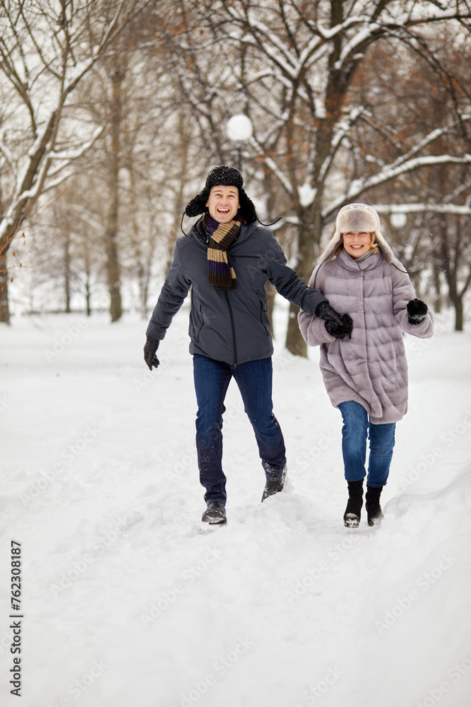 Smiling man and woman run holding hands in winter snow-covered park.