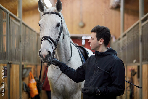 Fototapeta Naklejka Na Ścianę i Meble -  Man holds white horse by bridle in riding stables.