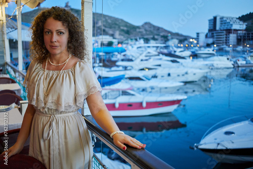 Smiling curly woman stands leaning on chair back and railing in floating cafe. © Pavel Losevsky