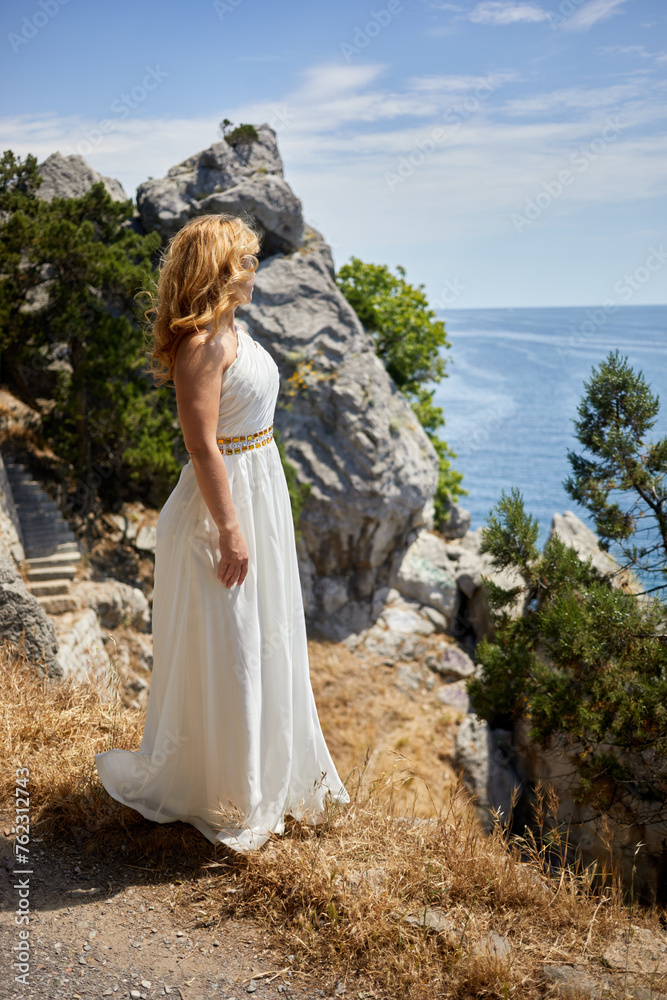 Blond woman in white dress with golden waist belt stands on cliff looking at sea on sunny day.