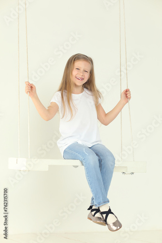 Happy little girl in jeans and white t-shirt swings in white studio and smiles