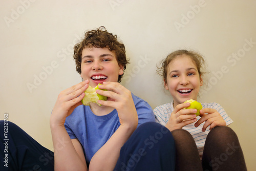 Happy brother and sister eat green apples and laugh near wall photo