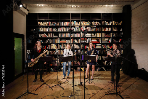 Four young pretty women play saxophone in room with bookshelves photo