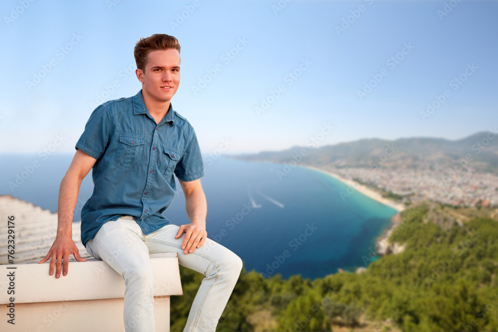 Smiling man in denim clothes sits on edge of building roof near sea shore