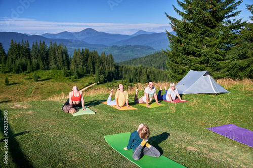 Group of people doing yoga pose outdoor in camping in the mountains. Adults and children on yoga mats, each doing a yoga pose under a clear blue sky in the morning. Young boy is instructor. photo